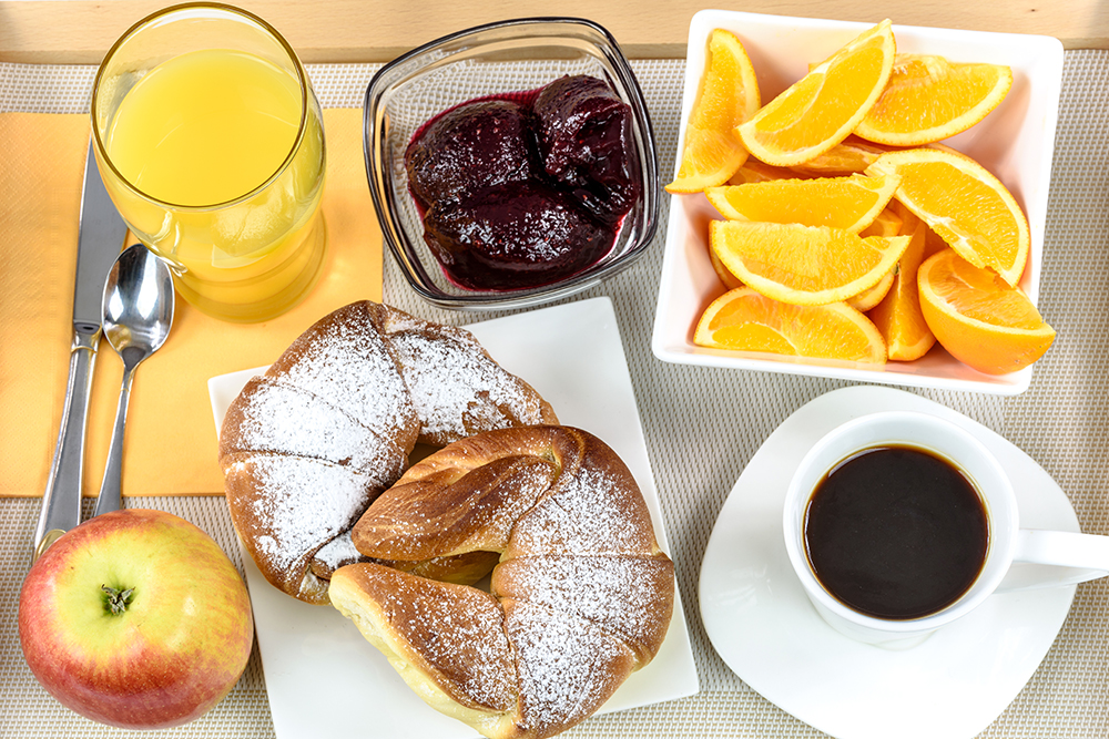 Overhead shot of a breakfast tray with croissants, coffee, oranges, juice, jam, apple and cutlery