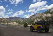 A yellow Jeep Wrangler drives in front of rocky mountains and an open sky